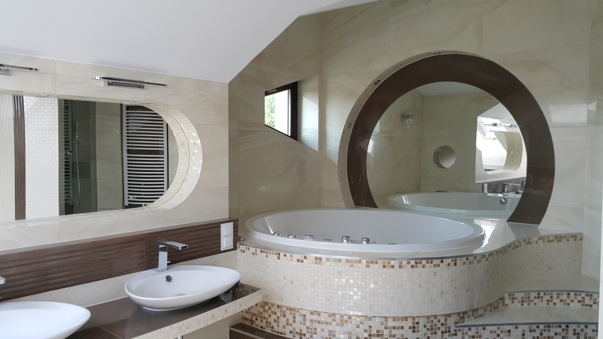 Jasna łazienka z wanną jacuzzi obudowaną mozaiką, owalne lustra z podświetleniem oraz umywalki nablatowe.