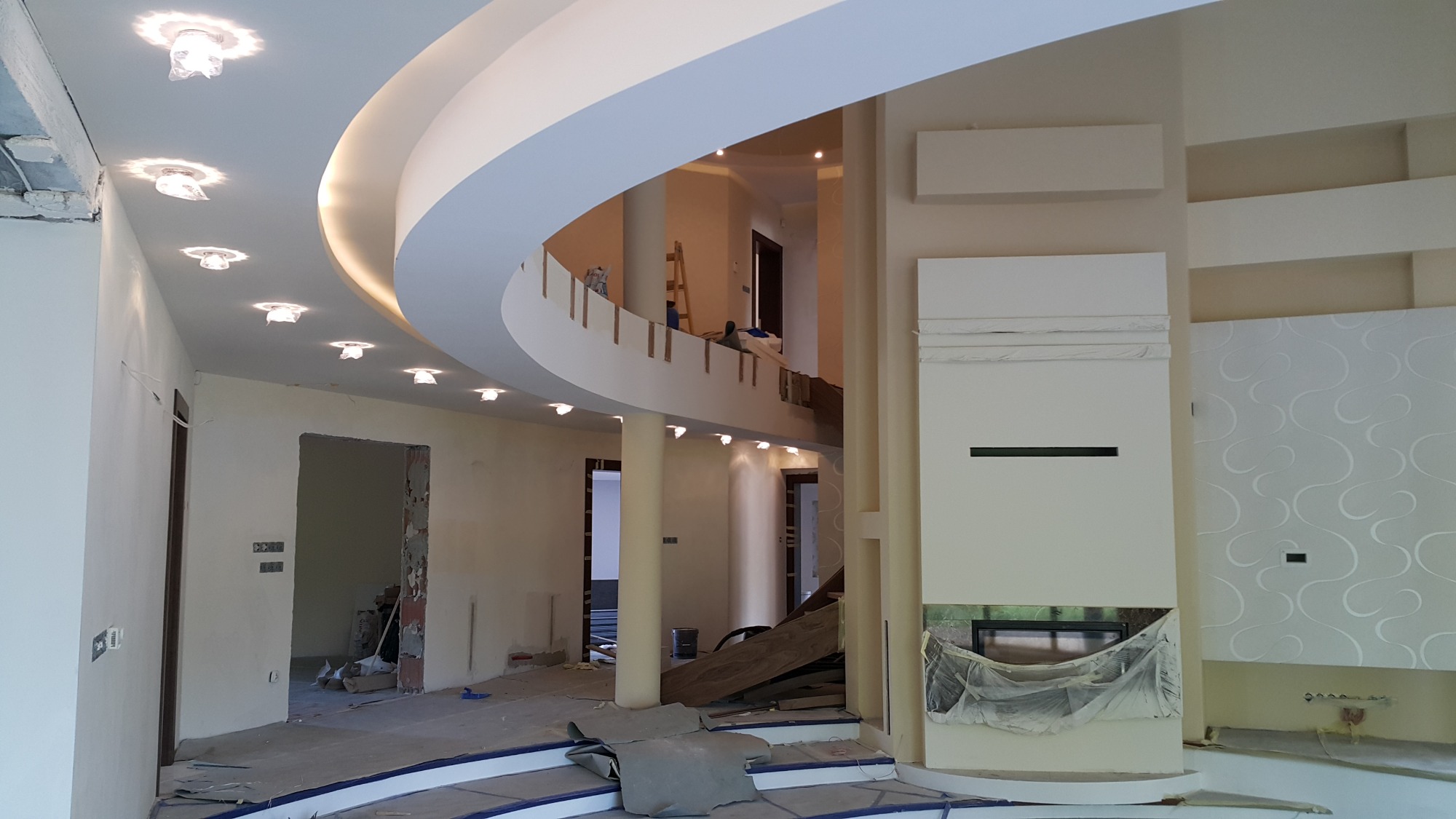 Wnętrze domu w trakcie prac wykończeniowych: widoczna dwupoziomowa przestrzeń z zaokrąglonym sufitem podwieszanym i oświetleniem punktowym, ściany w jasnych odcieniach, przygotowane do malowania...