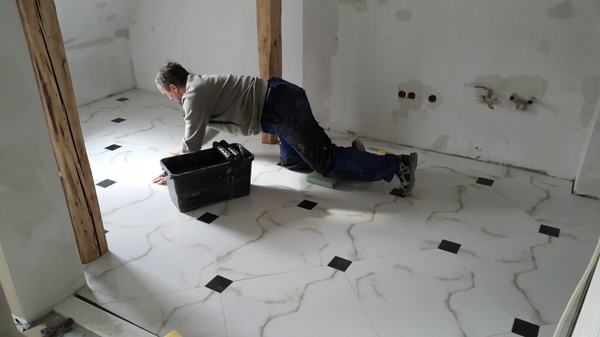 Mężczyzna w roboczym ubraniu układa białe płytki podłogowe z czarnymi wstawkami w pomieszczeniu z surowymi, drewnianymi elementami konstrukcyjnymi i niezamaskowanymi instalacjami wodnymi na ścianie.