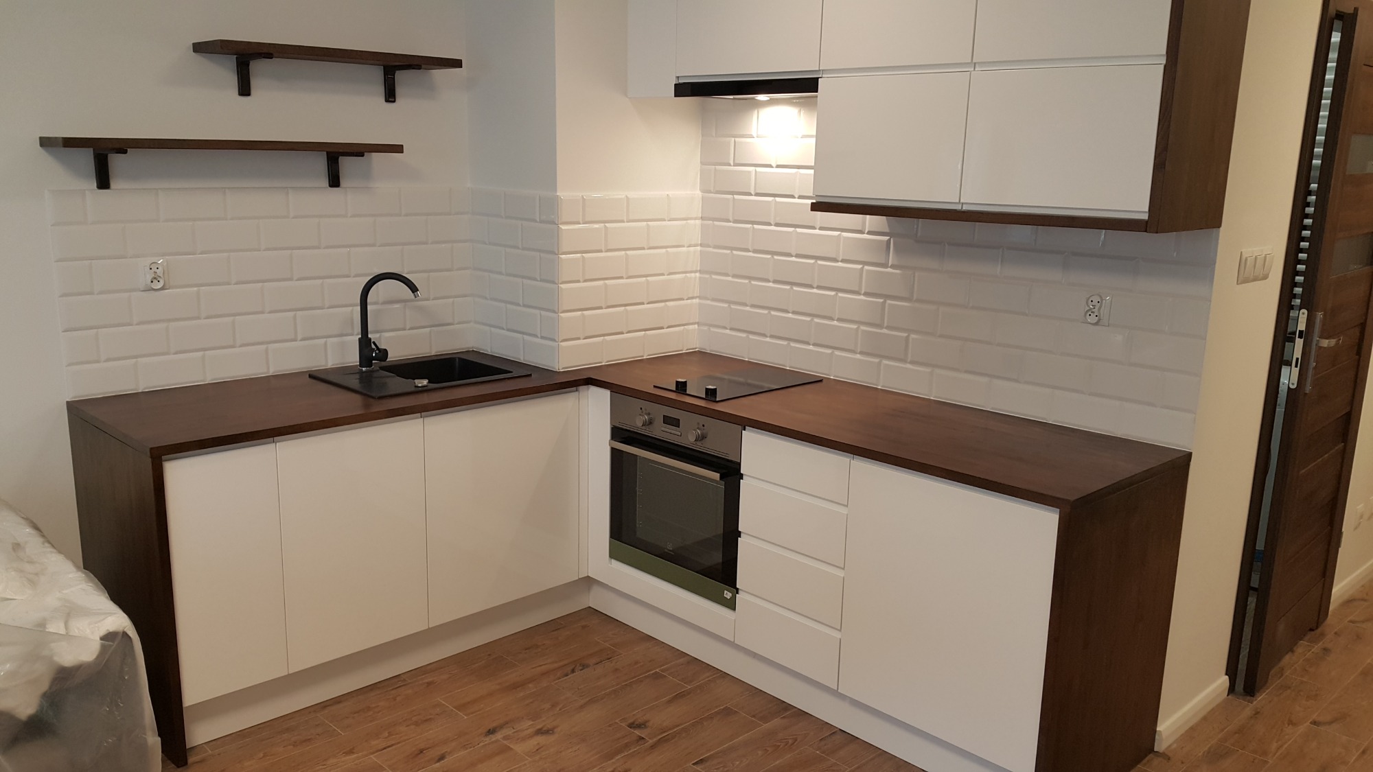 Nowoczesna kuchnia z białymi szafkami, blatem w kolorze ciemnego drewna i białymi płytkami typu metro, uzupełniona o czarny zlew i baterię oraz drewniane półki ścienne.