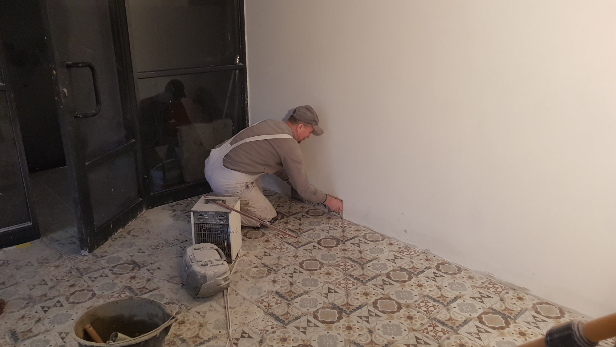 Mężczyzna w kombinezonie i czapce, na kolanach, wykańcza fugi przy podłodze z wzorzystych płytek ceramicznych, przy ścianie, widoczny grzejnik elektryczny i wiadro z narzędziami.