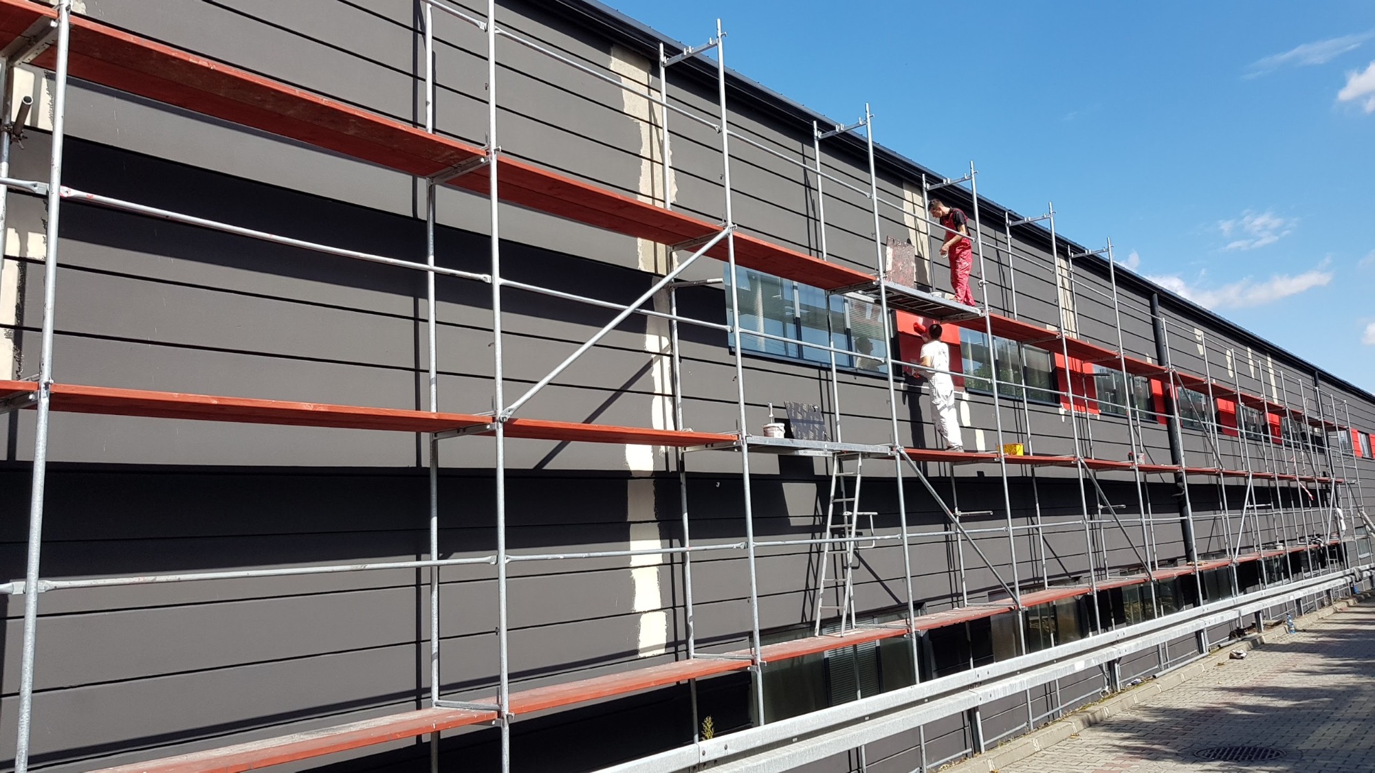 Malowanie elewacji budynku z rusztowania: dwóch malarzy w kombinezonach, jeden w czerwonym, drugi w białym, aplikują farbę na szarą elewację z poziomymi panelami, widoczne okna i fragment chodnika.
