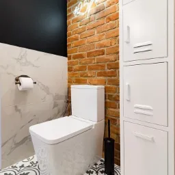Nowoczesna toaleta z białą ceramiką, wzorzystą podłogą, ścianą z czerwonej cegły i neonowym napisem, obok biała szafka w stylu industrialnym.