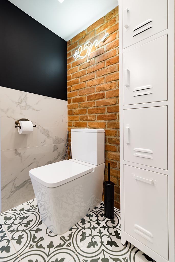 Nowoczesna toaleta z białą ceramiką, wzorzystą podłogą, ścianą z czerwonej cegły i neonowym napisem, obok biała szafka w stylu industrialnym.
