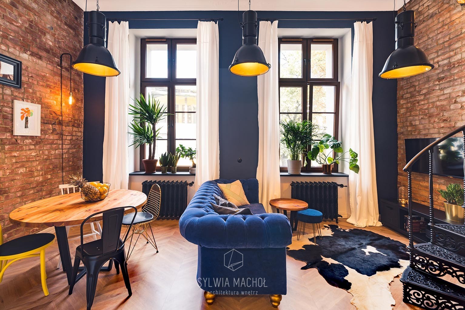 Salon w stylu industrialnym z ceglaną ścianą, granatowymi ścianami, drewnianym stołem, niebieską sofą i czarnymi lampami wiszącymi.