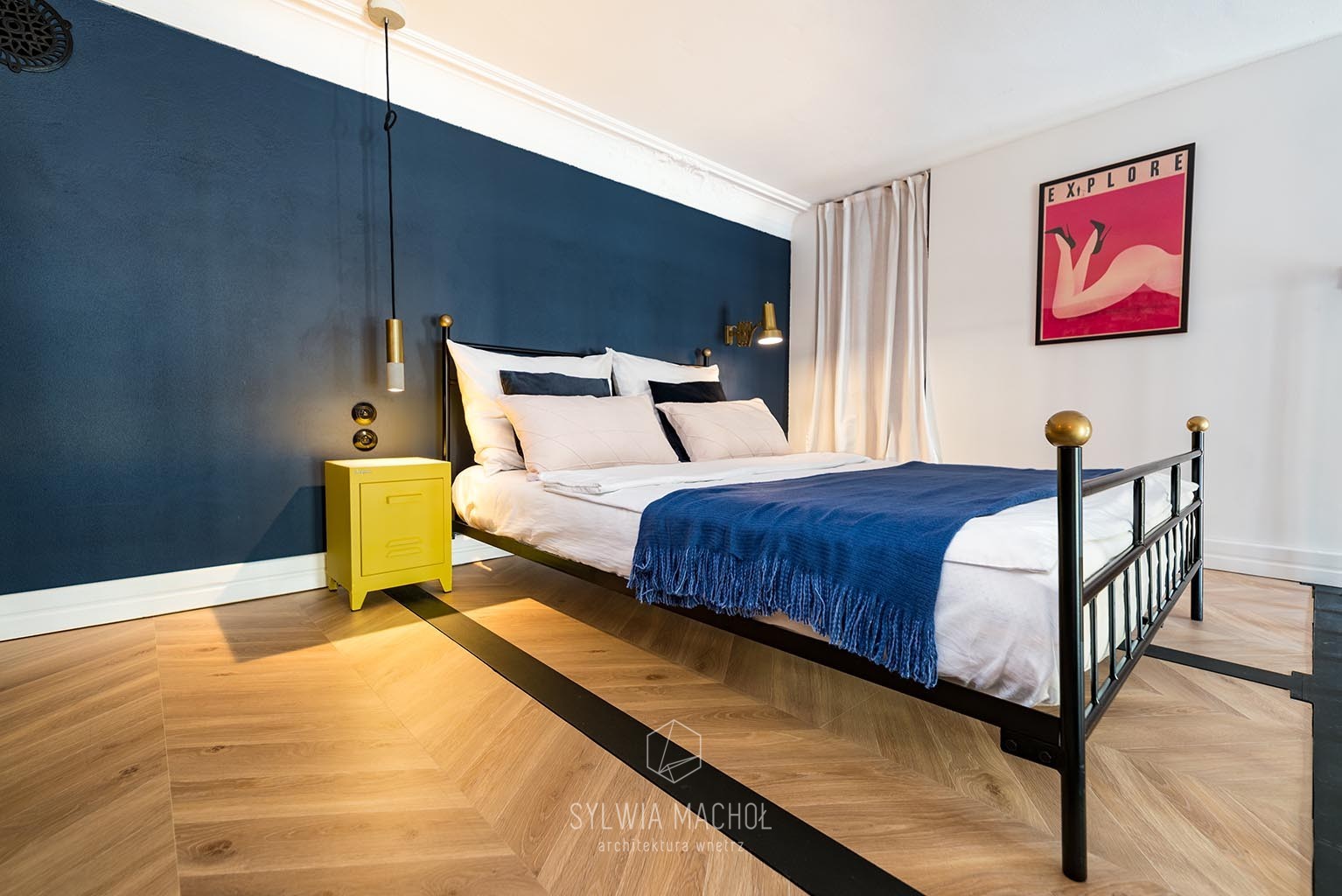 Sypialnia z ciemnoniebieską ścianą, parkietem w jodełkę z czarnymi paskami, czarnym łóżkiem z złotymi kulami, żółtą szafką nocną i plakatem na ścianie.