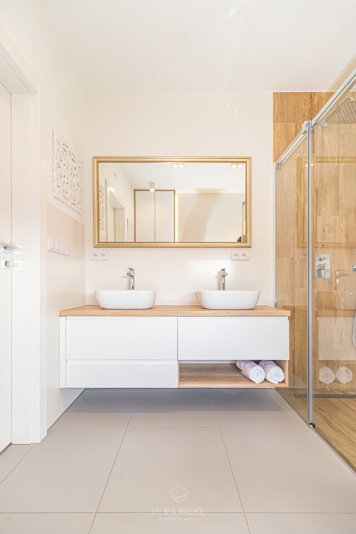 Jasna łazienka z dwoma umywalkami nablatowymi, złotym lustrem i kabiną prysznicową wykończoną drewnem, ręczniki w otwartej półce pod blatem.