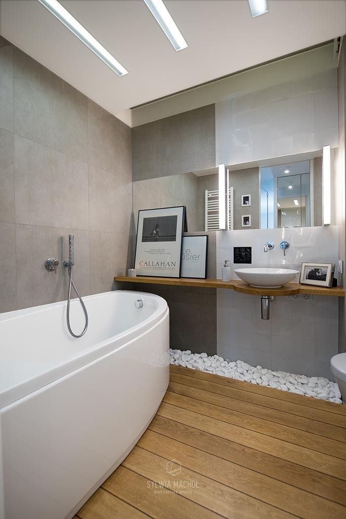 Nowoczesna łazienka z drewnianą podłogą, owalną wanną i umywalką na drewnianym blacie, dekorowana białymi kamieniami i plakatami w czarnych ramkach.