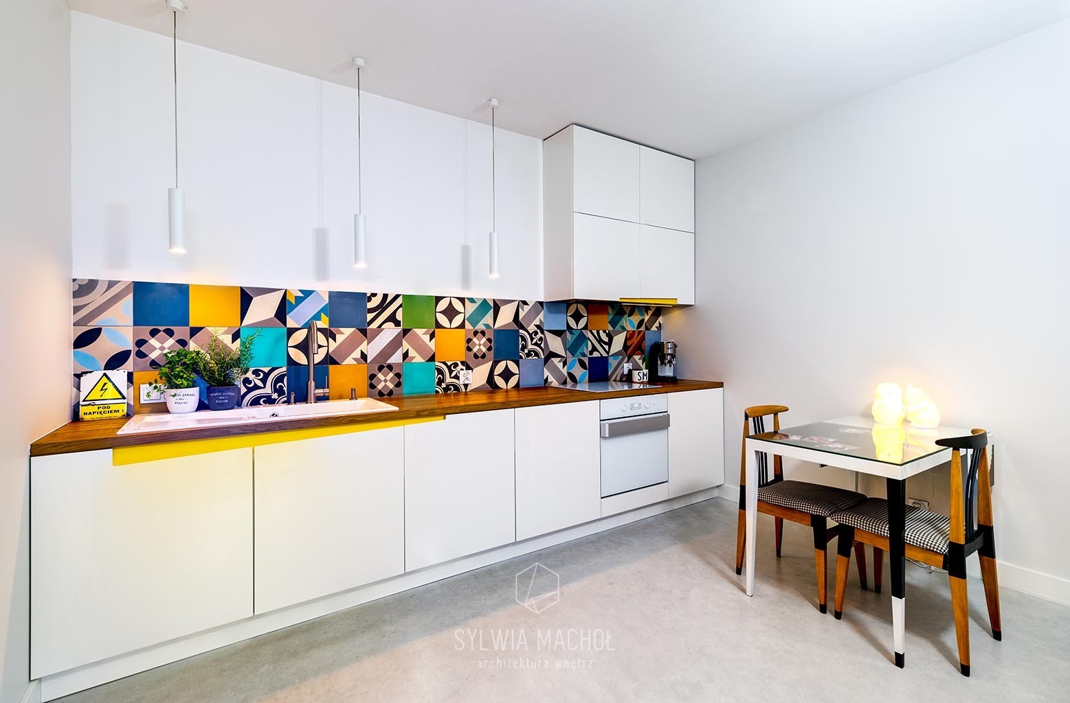Biała kuchnia z kolorowymi, wzorzystymi płytkami nad blatem, drewniany blat, białe szafki, stół z szklanym blatem i oryginalnymi krzesłami.