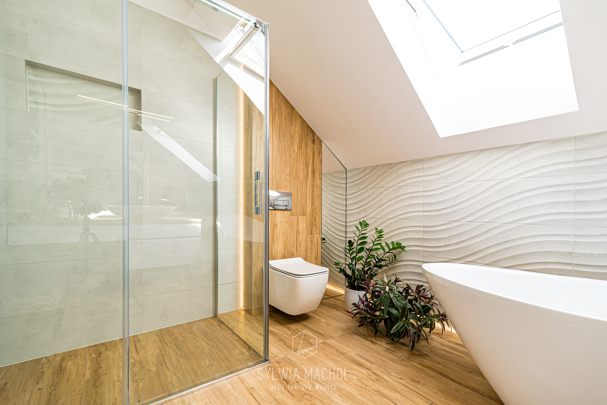 Nowoczesna łazienka na poddaszu z kabiną prysznicową, podwieszaną toaletą, wolnostojącą wanną i roślinami doniczkowymi, oświetlona światłem dziennym z okna dachowego.