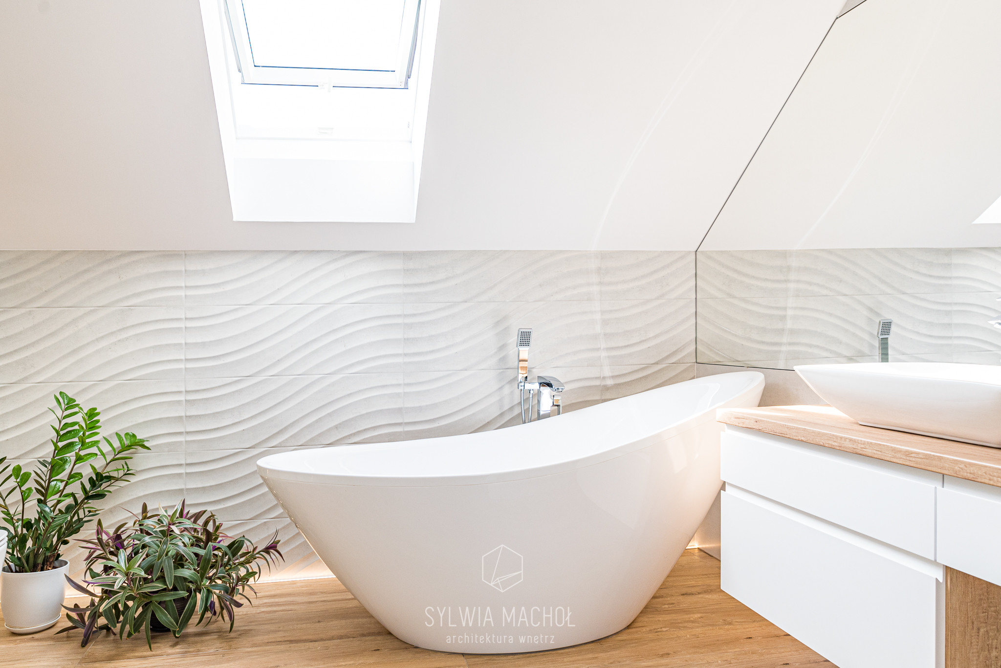 Nowoczesna łazienka na poddaszu z wolnostojącą białą wanną, umywalką nablatową na drewnianym blacie i roślinami w doniczkach, oświetlona światłem z okna dachowego.