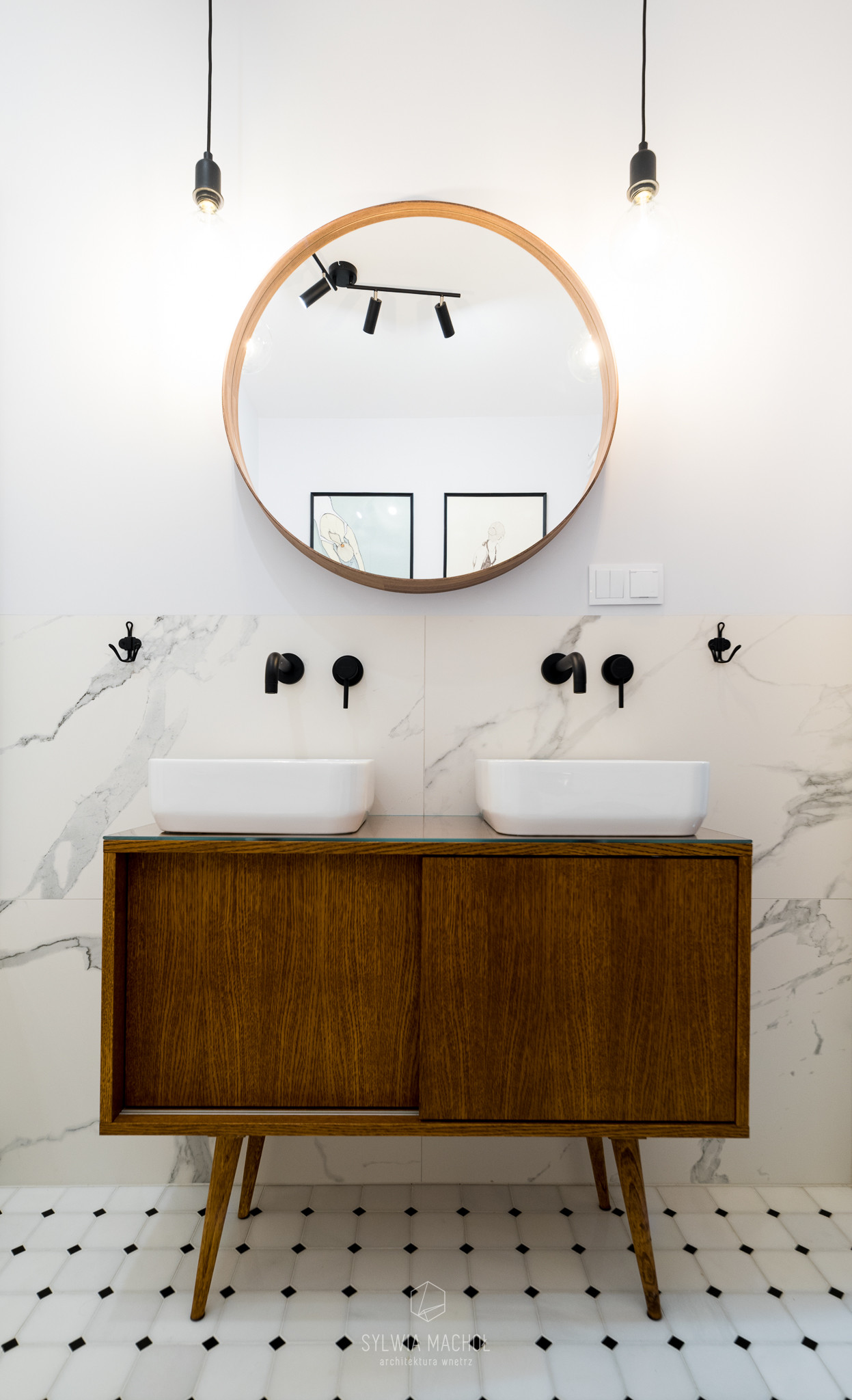 Łazienka z dwoma umywalkami nablatowymi, drewnianą szafką w stylu retro na nóżkach, okrągłym lustrem w drewnianej ramie i marmurowymi płytkami na ścianie oraz podłodze w biało-czarną szachownicę.