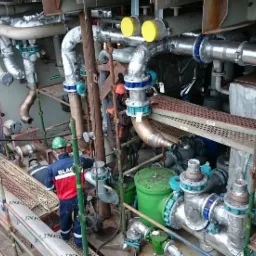 Prace instalacyjne w Tczewie: skomplikowany system rur i zaworów, częściowo zaizolowanych, widoczny pracownik w kasku na rusztowaniu.