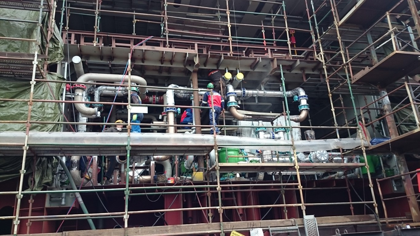 Prace instalacyjne rurociągów przemysłowych na rusztowaniu, widoczni pracownicy w kaskach i odzieży roboczej, fragment konstrukcji budynku w tle.