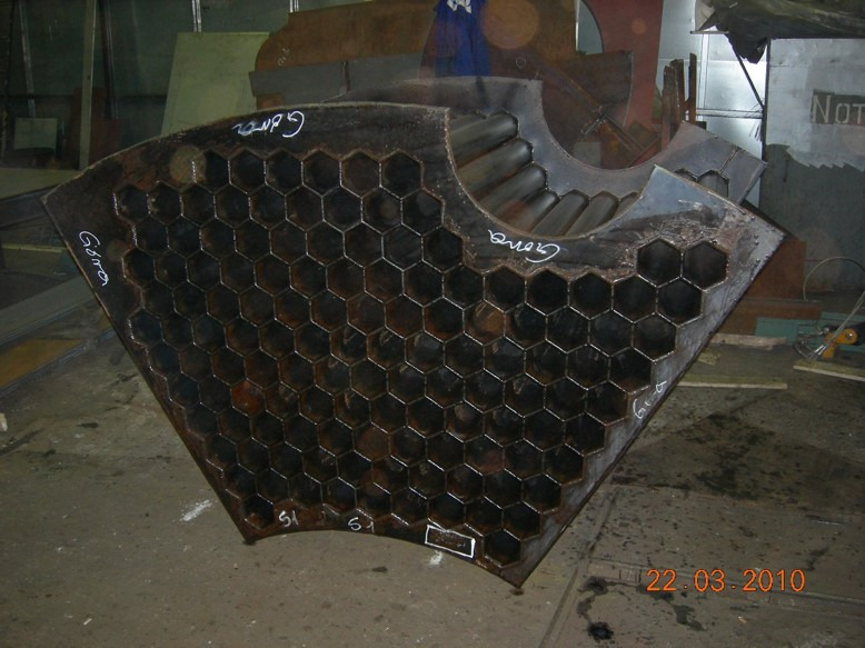 Metalowa konstrukcja w kształcie wycinka koła z otworami w kształcie sześciokątów, z widocznymi spawami i napisem 'Góra' wykonanym białą farbą, stojąca na betonowej posadzce w hali produkcyjnej.