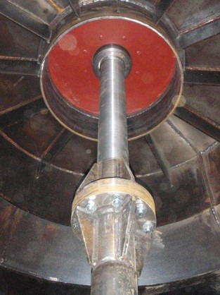 Wnętrze konstrukcji stalowej z centralnym, pionowym wałem podtrzymywanym przez wspornik z nakrętkami i śrubami, widoczna czerwona uszczelka wokół wału.