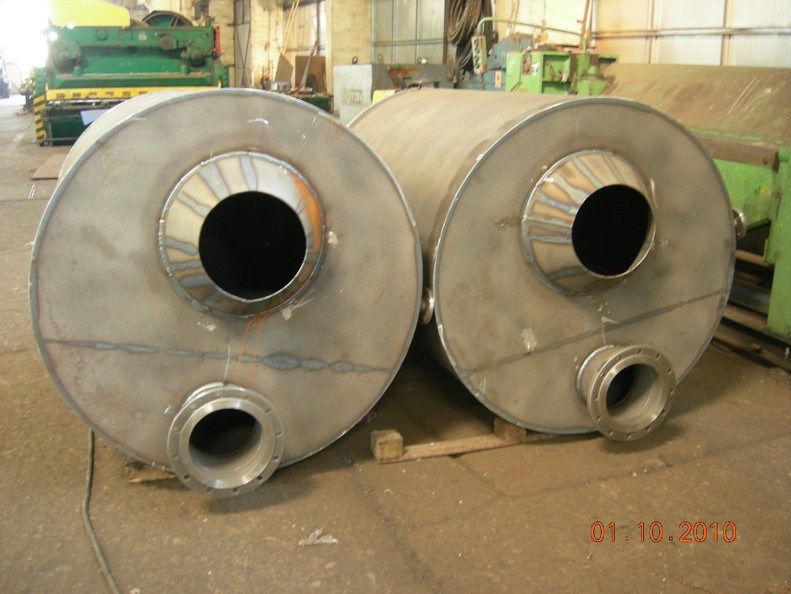 Dwa cylindryczne zbiorniki ze stali nierdzewnej z otworami, spawane, umieszczone na drewnianych paletach w hali produkcyjnej.