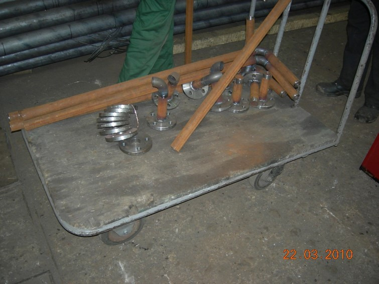 Wózek transportowy z rurami, kolankami z przyspawanymi kołnierzami i stos metalowych dysków w warsztacie.