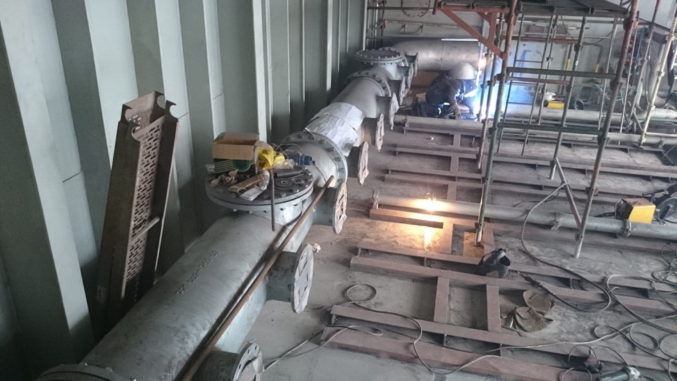 Spawanie rurociągu przemysłowego wewnątrz budynku, widoczne rusztowanie i narzędzia pracy.