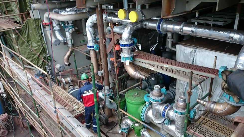 Prace instalacyjne w Tczewie: skomplikowany system rur i zaworów, częściowo zaizolowanych, widoczny pracownik w kasku na rusztowaniu.