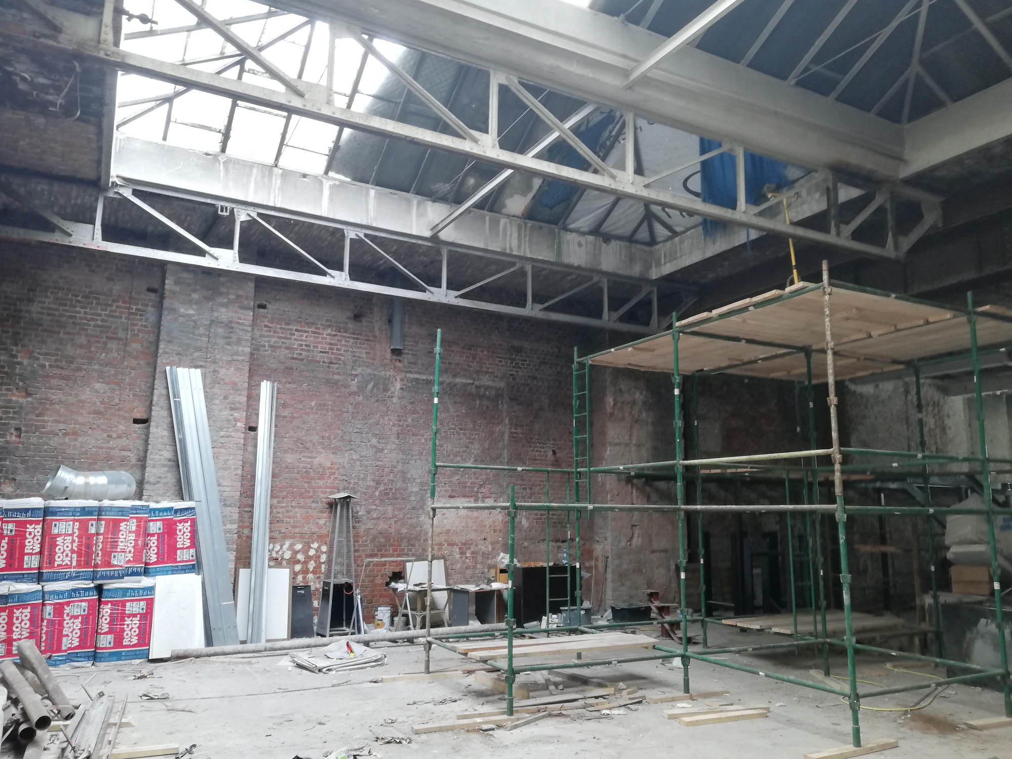 Wnętrze remontowanego budynku z odsłoniętą cegłą, widoczne rusztowania, materiały izolacyjne Rockwool, blachy i elementy konstrukcji dachu z oknami.