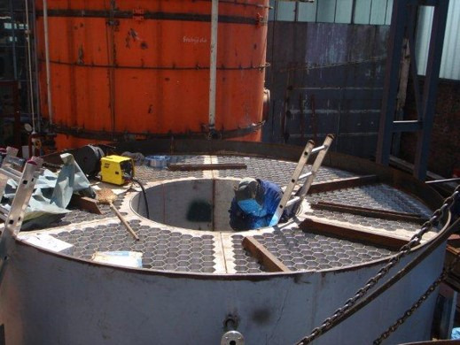 Spawacz w niebieskim kombinezonie i masce spawalniczej pracuje wewnątrz dużej metalowej konstrukcji z kratą o strukturze plastra miodu; obok drabina i spawarka, w tle pomarańczowy zbiornik.