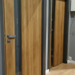 Dwie drewniane drzwi wewnętrzne z ościeżnicami w odcieniach szarości, jedna z metalową klamką, widoczne na tle szarej ściany i podłogi imitującej drewno.