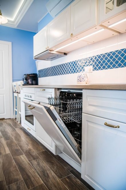 Biała kuchnia z otwartą zmywarką, niebieskie akcenty na ścianie i drewnopodobna podłoga, widoczny fragment ekspresu do kawy.