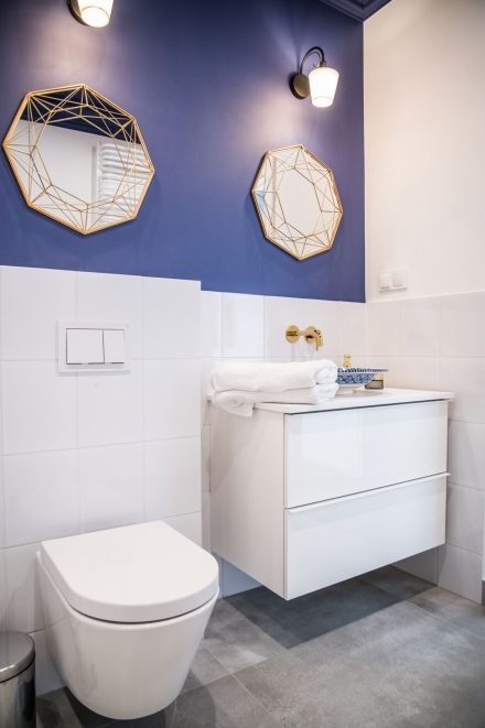 Nowoczesna łazienka z białymi płytkami, wiszącą toaletą, szafką z umywalką i dwoma geometrycznymi lustrami w złotych ramach na granatowej ścianie.