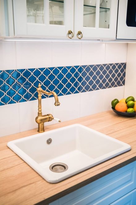 Biała, prostokątna umywalka kuchenna z baterią w kolorze starego złota, zamontowana na blacie z jasnego drewna. Nad umywalką pas niebieskich płytek z białym wzorem, a powyżej białe szafki...