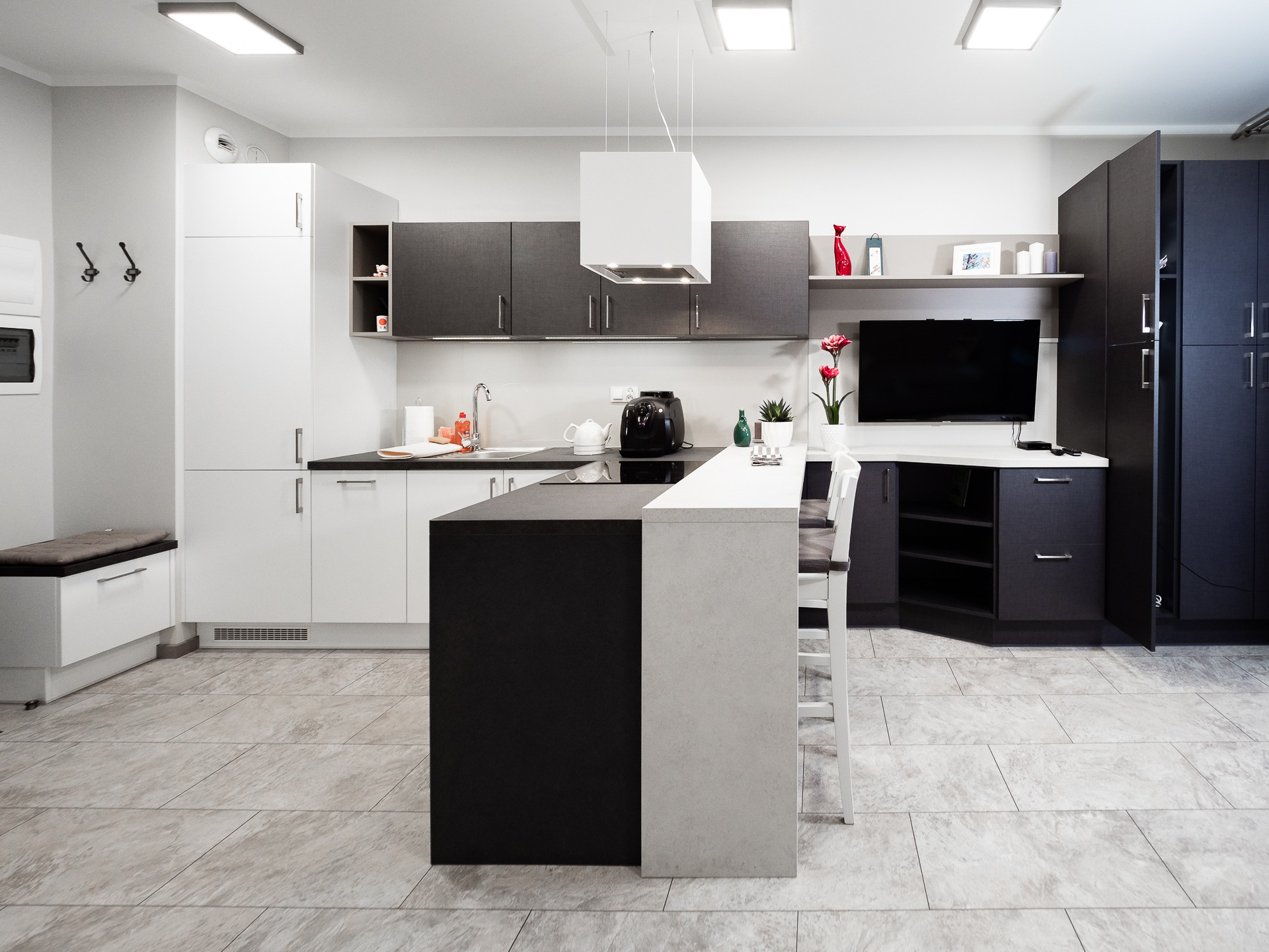 Nowoczesna kuchnia z wyspą, biało-czarne szafki, płytki podłogowe imitujące kamień, minimalistyczny okap i telewizor w zabudowie.