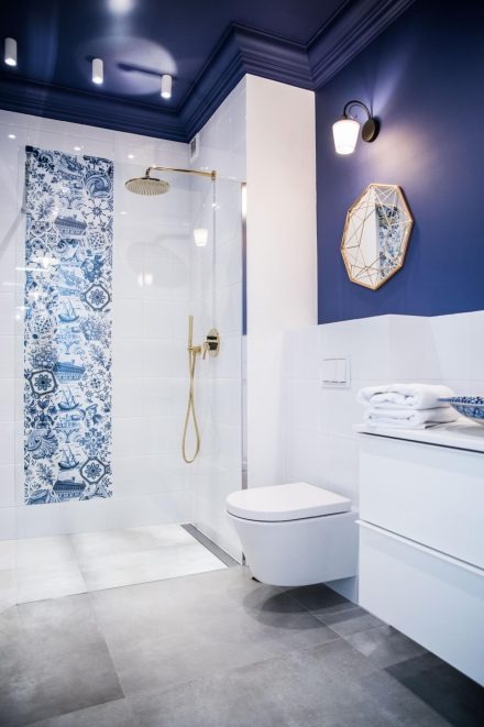 Nowoczesna łazienka z kabiną prysznicową typu walk-in, wykończona białymi płytkami z niebieskim, dekoracyjnym pasem płytek z motywami marynistycznymi, złotą armaturą, szarą podłogą i toaletą...