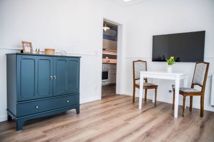 Jasne wnętrze po remoncie: ciemnoniebieska komoda z dekoracjami, biały stół z krzesłami i telewizorem na ścianie, widok na kuchnię w tle.