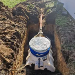 Montaż przydomowej oczyszczalni ścieków w wykopie, widoczna biała konstrukcja z niebieskimi rurami, przyłącze rury kanalizacyjnej, ziemia wokół wykopu.