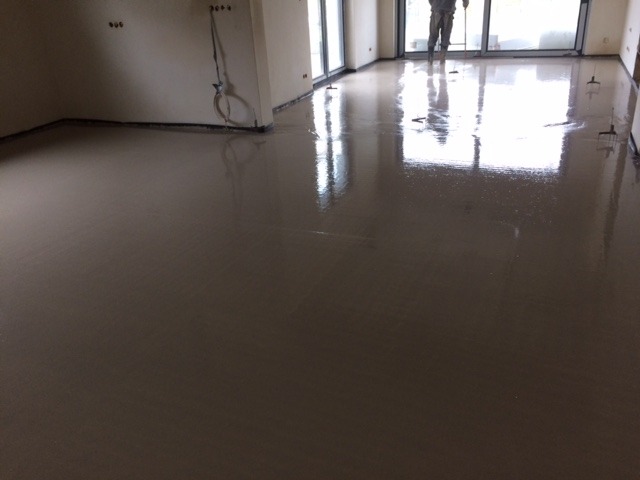 Świeżo wylana, mokra posadzka jastrychowa w przestronnym, jasnym pomieszczeniu z dużymi oknami balkonowymi, widoczny pracownik zacierający powierzchnię.