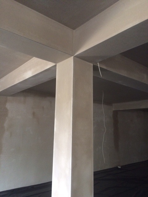 Wnętrze budynku w trakcie prac wykończeniowych, widoczne świeżo otynkowane ściany, słupy i strop. Podłoga zabezpieczona czarną folią.