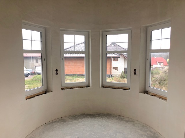 Wnętrze okrągłej wieżyczki z czterema białymi oknami, świeżo otynkowane ściany w kolorze złamanej bieli, surowa posadzka, widok na domy przez okna.