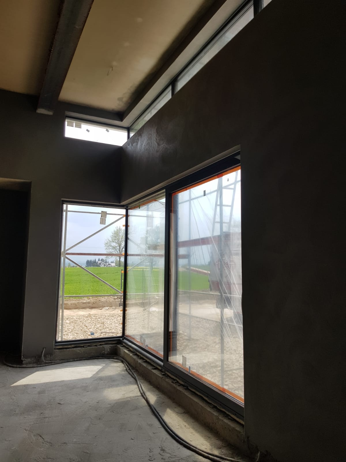 Wnętrze w trakcie prac wykończeniowych: surowa posadzka, świeżo otynkowane szare ściany z dużymi oknami, widoczne rusztowanie za folią ochronną okienną, oraz przewody na podłodze.