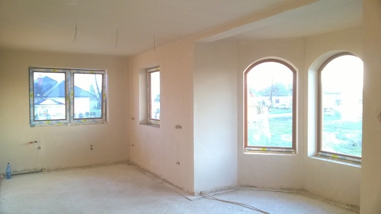 Wnętrze w stanie deweloperskim po nałożeniu jasnego tynku, widoczne dwa okna łukowe i jedno prostokątne, wystające kable i rurki instalacyjne.