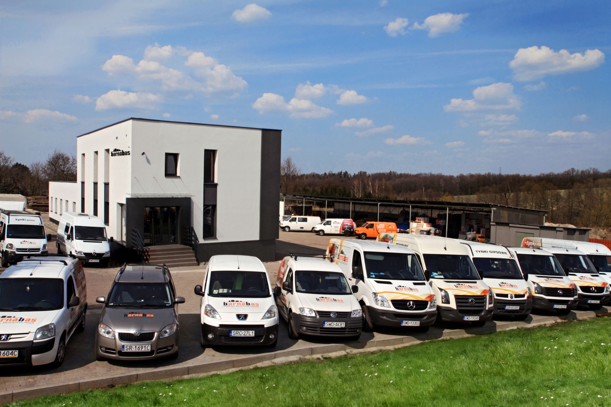 Flota samochodów firmowych z logo 'barnabas' zaparkowana przed nowoczesnym, dwukondygnacyjnym budynkiem biurowym w słoneczny dzień. Na niektórych autach widoczne napisy 'tynki gipsowe'.