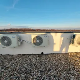 Trzy klimatyzatory marki AUX zamontowane na białej ścianie z widocznym cieniem człowieka, tło to kamienny dach i błękitne niebo z drzewami w oddali.