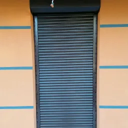 Zamknięta, brązowa osłona okienna z widocznymi kablami elektrycznymi u góry, zamontowana na pomarańczowej elewacji z niebieskimi pasami.