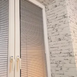 Zamknięte i częściowo otwarte poziome żaluzje aluminiowe w oknie PCV, na tle ściany z białej cegły.