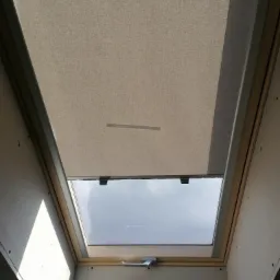 Beżowa, częściowo zasłonięta roleta w oknie dachowym, widok z dołu na jasne niebo