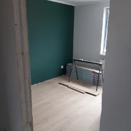 Pokój po położeniu paneli podłogowych w jodełkę, ściana pomalowana na ciemny zielony kolor, widoczna sterta paneli na stojaku i surowa ościeżnica drzwi.