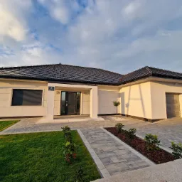 Nowoczesny, parterowy dom z białymi ścianami i ciemną dachówką, numer domu '7' nad wejściem, otoczony trawnikiem i ścieżką z kostki brukowej w promieniach zachodzącego słońca.