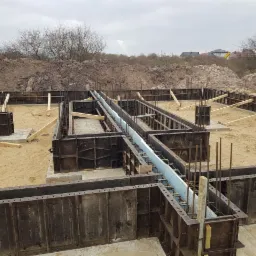 Szczegółowy widok wykopu fundamentowego z szalunkami stalowymi, zbrojeniami i izolacją termiczną, przygotowanego do wylania betonu, z widoczną stertą ziemi w tle.