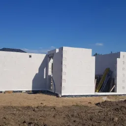 Wznoszone ściany parterowego budynku z białych bloczków betonu komórkowego na fundamencie, widoczne drewniane stemple i niebieski przewód instalacyjny w otworze okiennym.