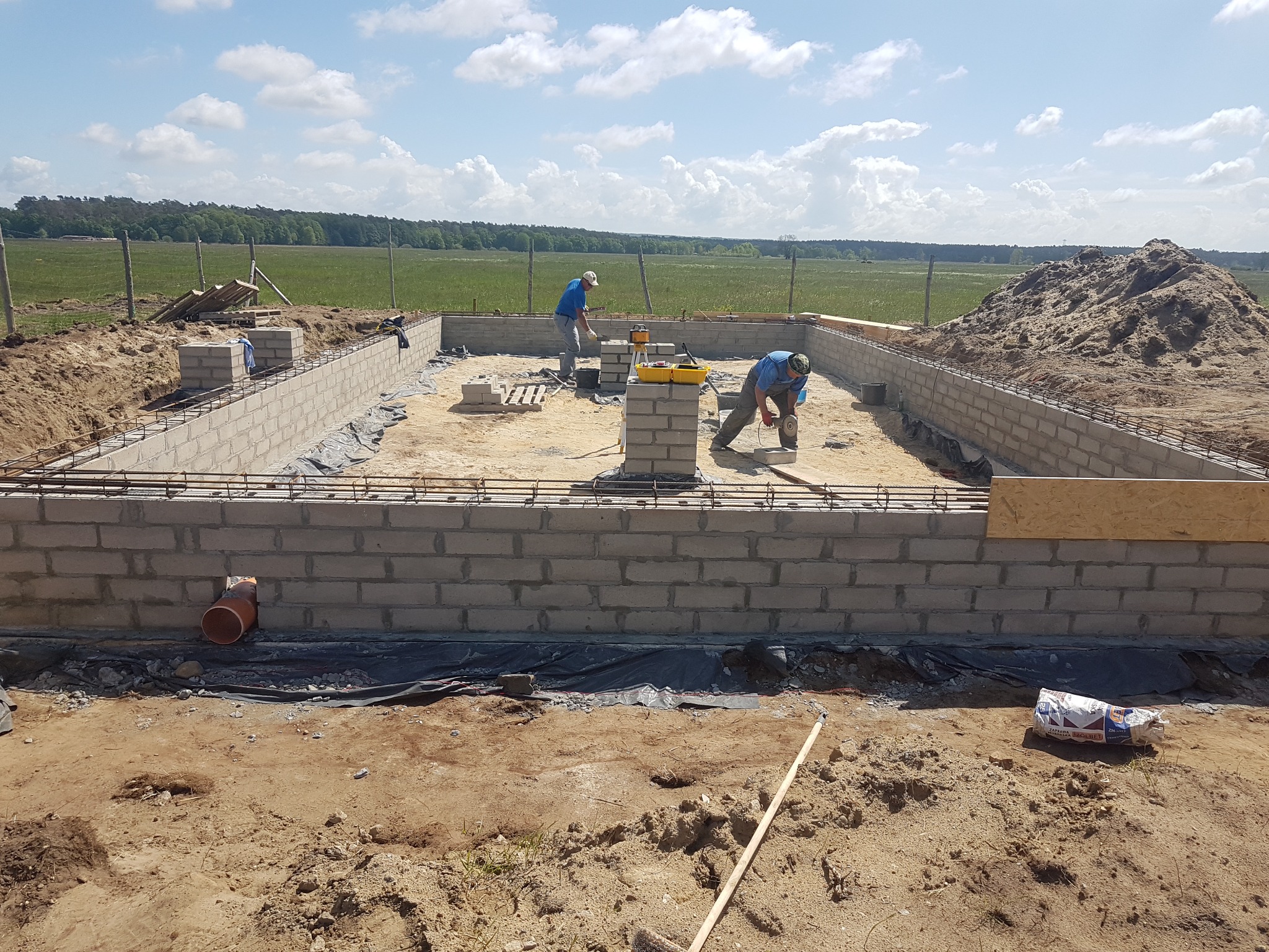 Budowa fundamentów pod dom jednorodzinny w Szczecinie: dwóch pracowników w trakcie murowania ścian z bloczków betonowych, widoczne zbrojenia, poziomica laserowa i narzędzia.