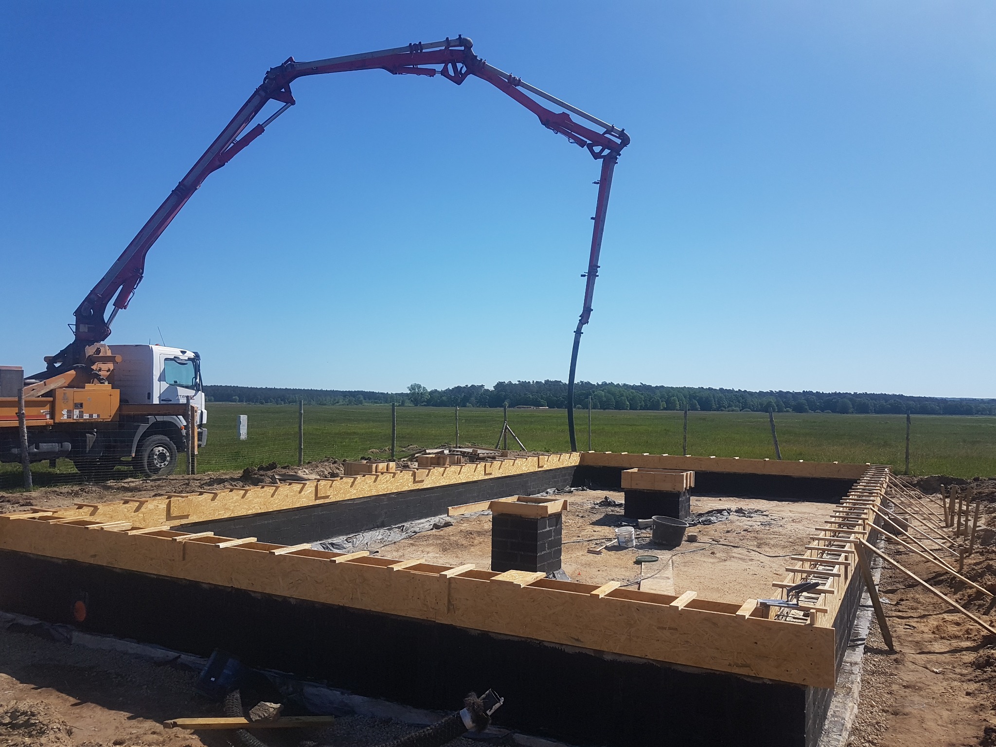 Wylewanie betonu do szalunku fundamentu domu jednorodzinnego za pomocą pompy do betonu na tle zielonej łąki i błękitnego nieba.